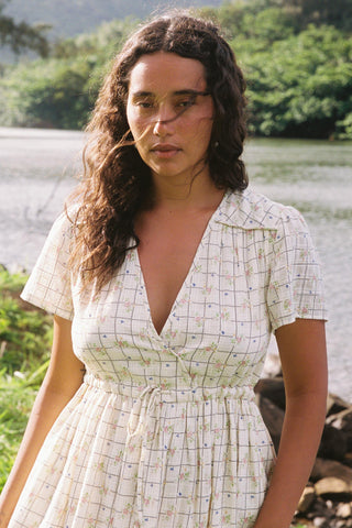 Woman in a cream checkered dress standing by a body of water with greenery in the background