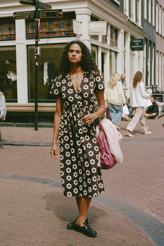 A woman stands on a city sidewalk wearing a black midi dress with a white floral pattern, holding a pink bag. The background shows a street scene with buildings and other pedestrians.