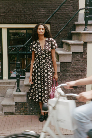 A woman wearing a brown midi dress with a white floral pattern stands on stone steps in front of a brick building, holding a pink handbag. A person riding a white bicycle is partially visible in the foreground.