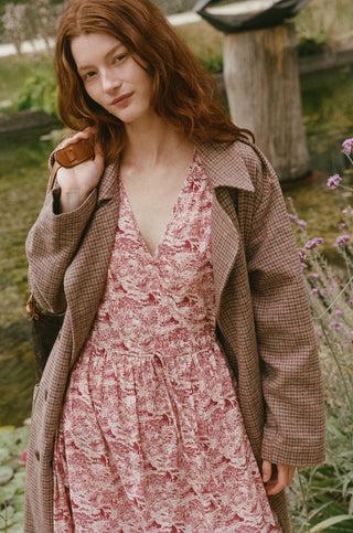 A woman wearing a pink and burgundy toile midi dress under a brown plaid coat, standing outdoors near a garden pond.