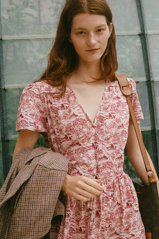 A woman wearing a pink and burgundy toile print wrap dress with short sleeves and a V-neckline, holding a brown plaid jacket and carrying a brown shoulder bag, set against a green glass-paneled background.