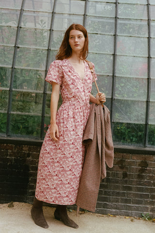 A woman stands in front of a greenhouse-style glass wall wearing a pink and burgundy toile midi dress with short sleeves and holding a brown plaid coat and a shoulder bag.