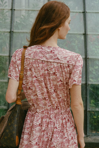 A woman viewed from behind wearing a short-sleeved pink and burgundy toile print dress, carrying a brown shoulder bag with a monogram pattern, standing in front of a glass-paneled wall.