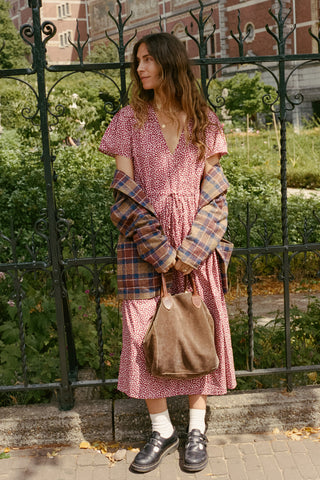 A woman stands outdoors in front of a black wrought-iron fence and greenery, wearing a pink and white patterned dress with a plaid blazer draped off her shoulders, white socks, black loafers, and carrying a brown tote bag.