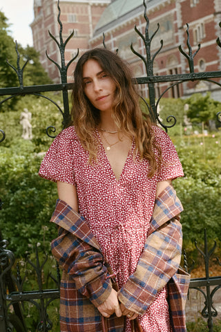 A woman stands outdoors in front of a black wrought-iron fence and greenery, wearing a red-and-white patterned wrap dress with short flutter sleeves and holding a plaid blazer draped off her shoulders.