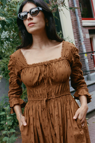 A woman wearing a brown smocked dress with ruffled sleeves and square neckline, styled with black sunglasses, standing outdoors in front of a brick building.
