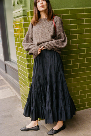 Woman wearing a brown sweater and black tiered skirt against a green tiled wall.