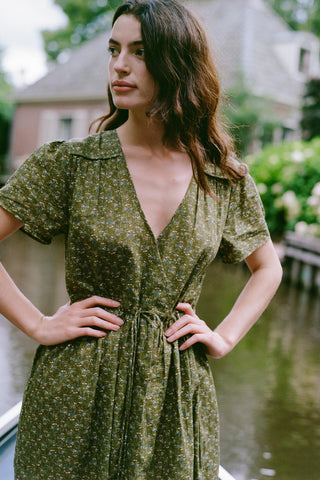 A woman wearing a short-sleeve olive green floral dress with a V-neck and waist tie, standing outdoors near water with a blurred house in the background.