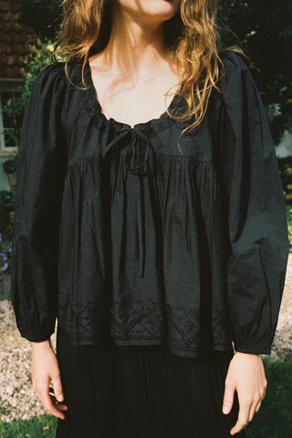A woman wearing a black long-sleeve blouse with a tie-neck detail and embroidered lace trim at the hem, photographed outdoors in natural light.