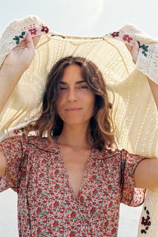 A woman wearing a red and green apple print short-sleeve dress stands holding a cream knit sweater over her head