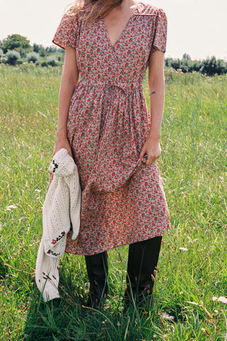 A woman wearing a red and green apple print short-sleeve dress stands in a grassy field 