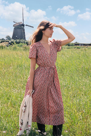 A woman wearing a red and green apple print short-sleeve dress stands in a grassy field with a windmill and cloudy sky in the background, holding a cream knit sweater.