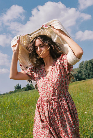 A woman wearing a red and green apple print short-sleeve dress stands in a grassy field, holding a cream knit sweater above her. The sky is blue with some clouds.